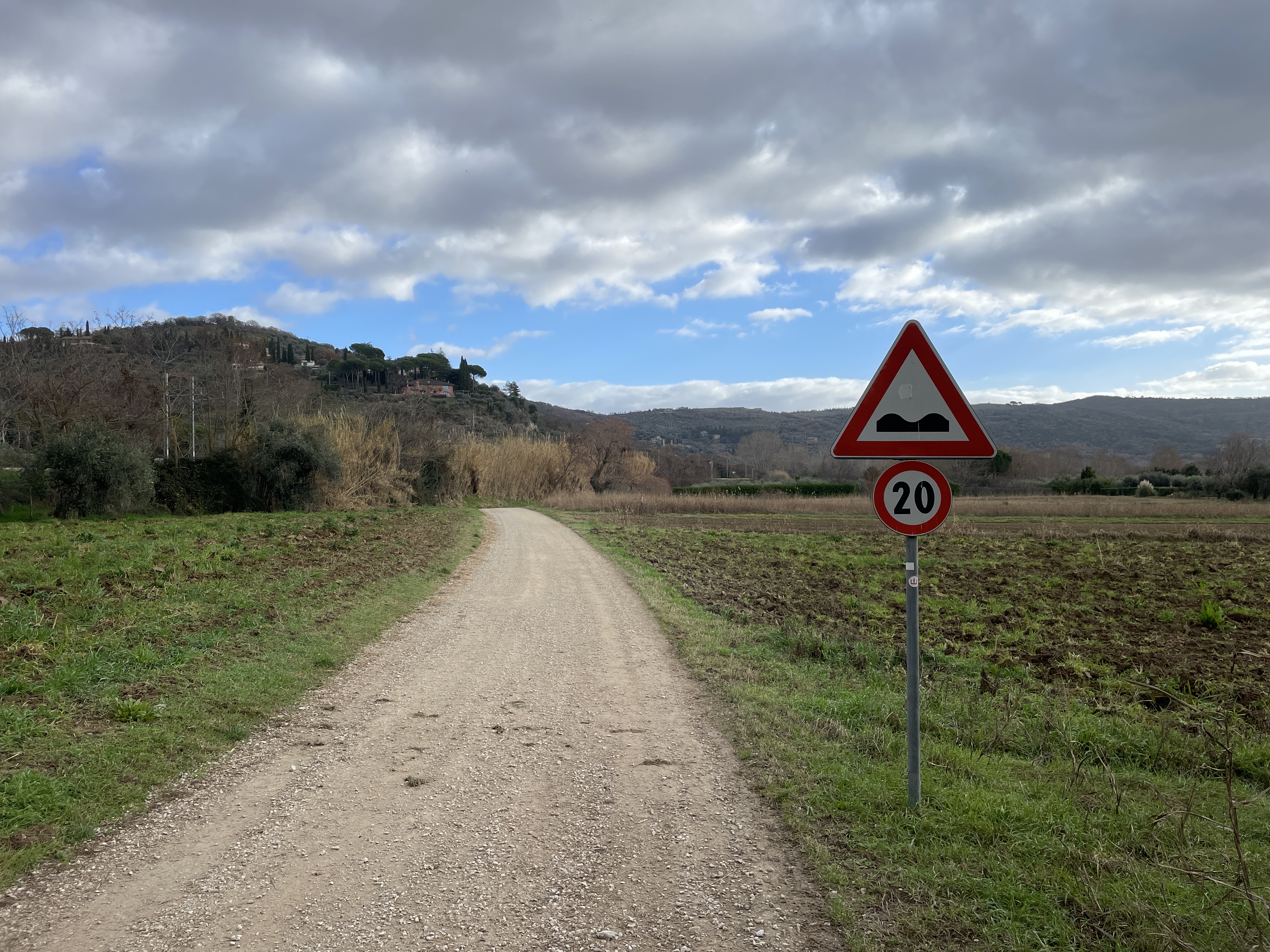 Section of cycle path with wide gravel track in the countryside, with signpost on the right for hump and 20 km/h limit.